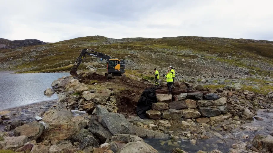 Tre personer på befaring ved gravemaskin på dam ved drikkevann på fjellet.