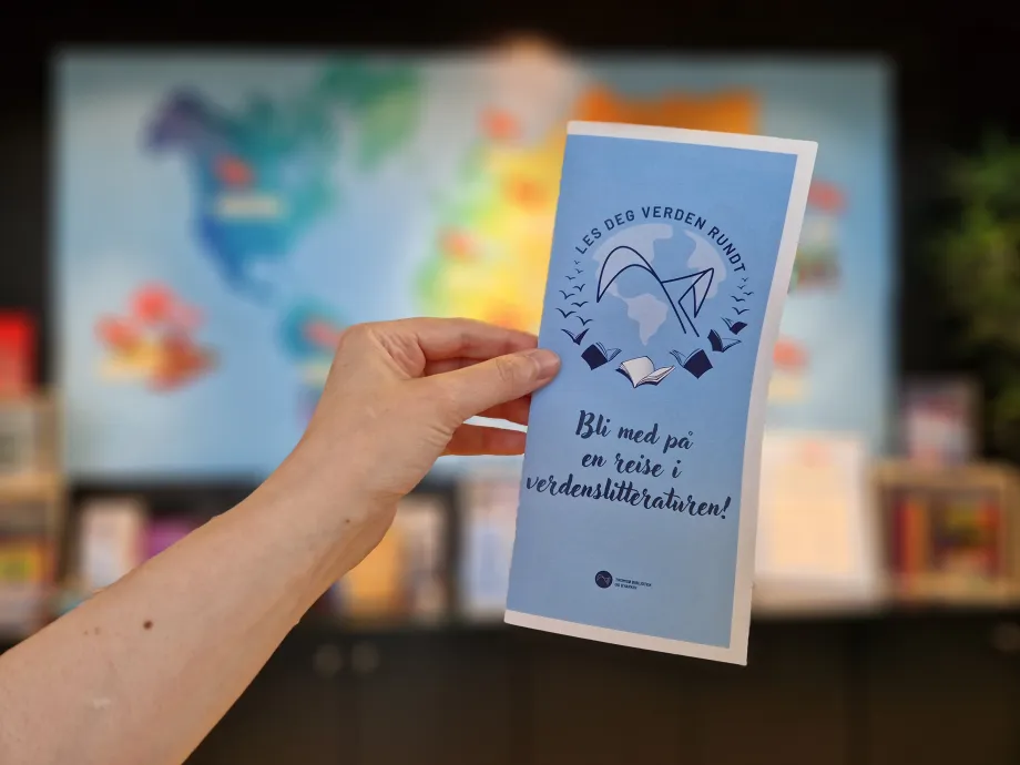 H&aring;nd holder en brosjyre med bl&aring; skrift p&aring; lysebl&aring; bakgrunn. Verdenskart i bakgrunnen. Foto