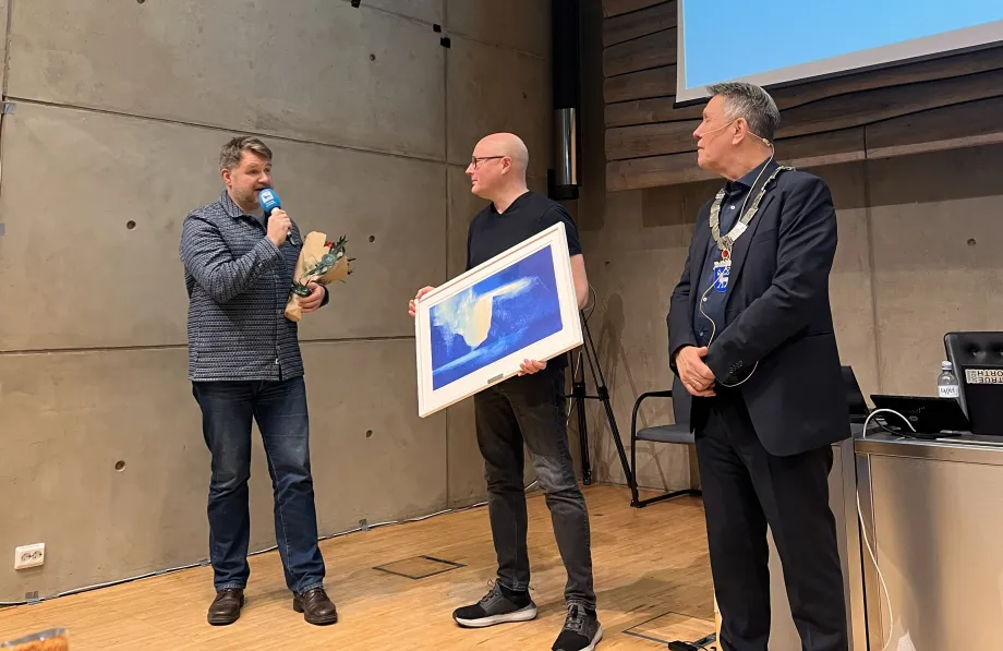 To medlemmer i Tromsø Storband står ved siden av ordføreren. Det ene bandmedlemmet holder mikrofon mot munnen. Kollegaen holder kunstverk.