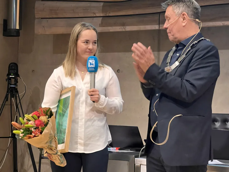 Vilde Nilsen står med blomsterbukett og kunstverk i hånda. Holder tale via mikrofon. Ordføreren står ved siden av og applauderer.