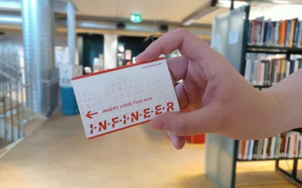 Et hvitt og oransje kort holdes av en h&aring;nd. I bakgrunnen st&aring;r det bokhyller. Foto.