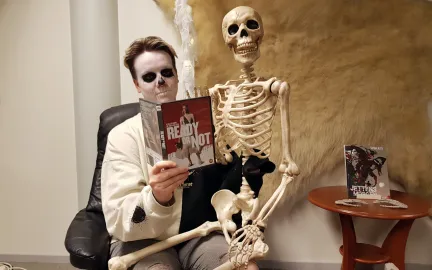 Kvinne med Halloweensminke i ansiktet sitter med et leke-skjelett p&aring; fanget. Hun holder opp et filmdeksel. Foto