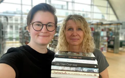 Kvinne med kort, brunt h&aring;r og runde, m&oslash;rke briller st&aring;r sammen med kvinne med skuldelangt, blondt kr&oslash;llet h&aring;r. De holder en stabel b&oslash;ker i hendene og smiler mot kamera. Foto.