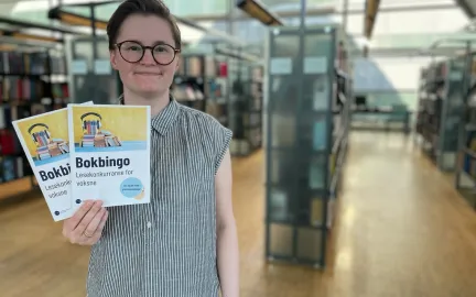 Kvinne med kort brunt h&aring;r, briller og gr&aring;-og-hvit-stripete skjorte holder to ark og smiler mot kamera. I bakgrunnen er vinduer og bokhyller. Foto.