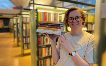 Kvinne med halvlangt, brunt h&aring;r og lys gr&aring; t-skjorte holder en bunke b&oslash;ker og smiler mot kamera. I bakgrunnen st&aring;r reoler med b&oslash;ker. Foto