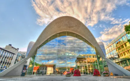 Bygning med glassfasade og hvelvede takpartier i hvitt. Bl&aring; himmel i bakgrunnen, overgangsfelt og blomsterdekorasjoner i forgrunnen. Foto