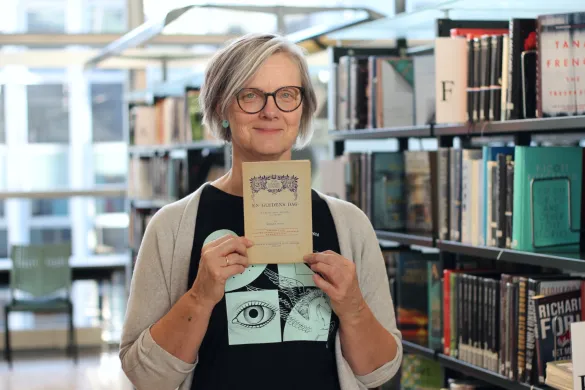 Kvinne med briller og gr&aring; cardigan holder opp en gammel bok og smiler mot kamera. Foto