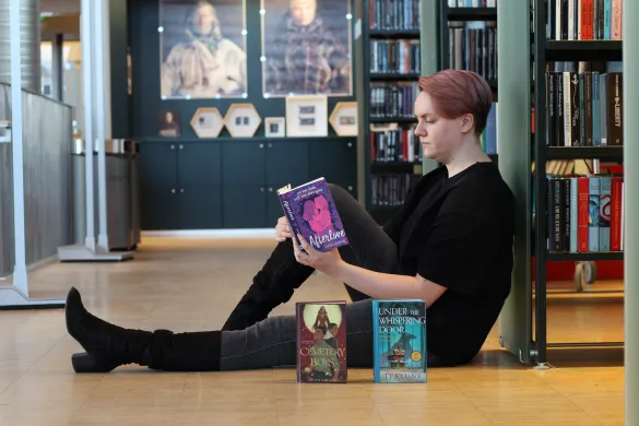 Regine_dødsforelska Kvinne sitter på gulvet og leser ei bok med ryggen lent mot ei bokhylle. Hun er kledd i svart og har lange, svarte støvletter på seg. Foran henne står to bøker med fronten mot kamera. Foto.