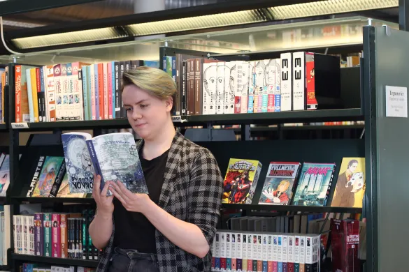 Kvinne i rutete skjorte st&aring;r lent mot en bokhylle og leser i ei bok. Foto.