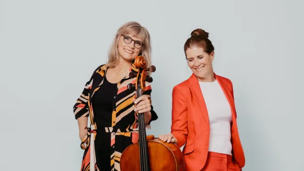 To damer ser i kamera. Den til venstre holder en cello. Foto.