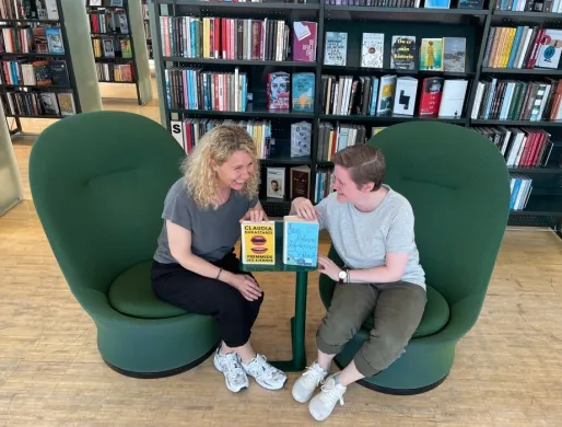 AnneBogAnja-sommerlesetips-2023 To kvinner ler mot hverandre og har to bøker stående mellom seg på et lite bord. De sitter i grønne lenestoler med bokhyller bak seg. Foto.