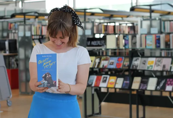 AnneK-sommerlesetips-2023 Kvinne i hvit t-skjorte og blått skjørt smiler og ser ned mot ei blå bok hun holder foran seg. I bakgrunnen er det bokhyller. Foto.