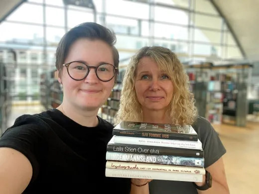 Kvinne med kort, brunt h&aring;r og runde, m&oslash;rke briller st&aring;r sammen med kvinne med skuldelangt, blondt kr&oslash;llet h&aring;r. De holder en stabel b&oslash;ker i hendene og smiler mot kamera. Foto.