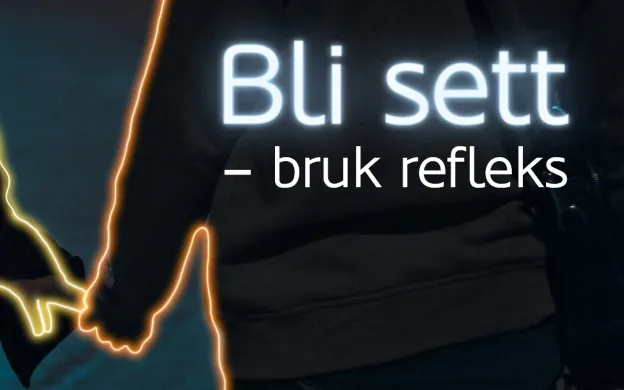 To personer som holder hender g&aring;r langs m&oslash;rk vei. Tekst i bildet sier: Bli sett - bruk refleks.