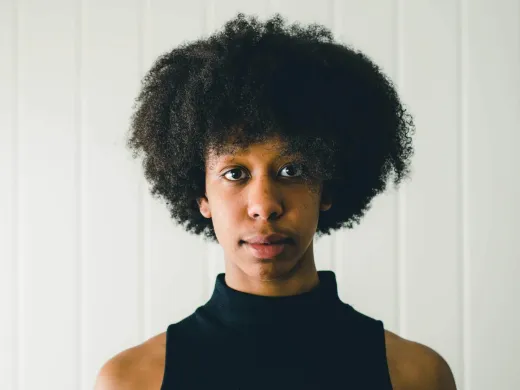 Kvinne med m&oslash;rkt, krusete h&aring;r og svart, kortermet topp ser inn i kamera. Foto.