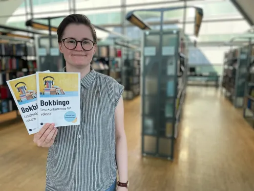 Lesetips til bokbingo Kvinne med kort brunt hår, briller og grå-og-hvit-stripete skjorte holder to ark og smiler mot kamera. I bakgrunnen er vinduer og bokhyller. Foto.