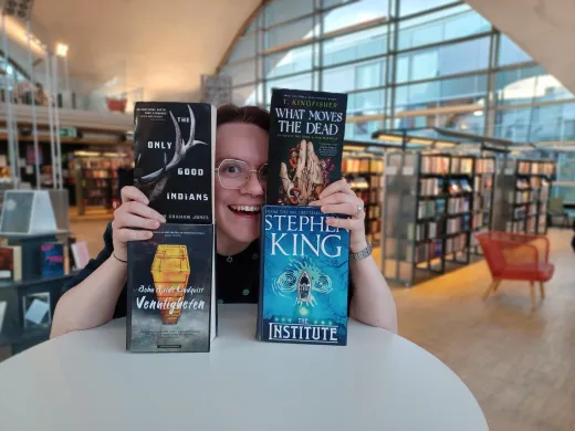 Oktober-lesetips 2024 Kvinne holder opp fire bøker og smiler bredd. Ansiktet hennes stikker frem mellom bøkene. I bakgrunnen en stor glassvegg og bokreoler. Foto.