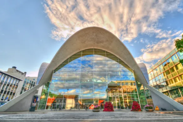 Bygning med glassfasade og hvelvede takpartier i hvitt. Bl&aring; himmel i bakgrunnen, overgangsfelt og blomsterdekorasjoner i forgrunnen. Foto